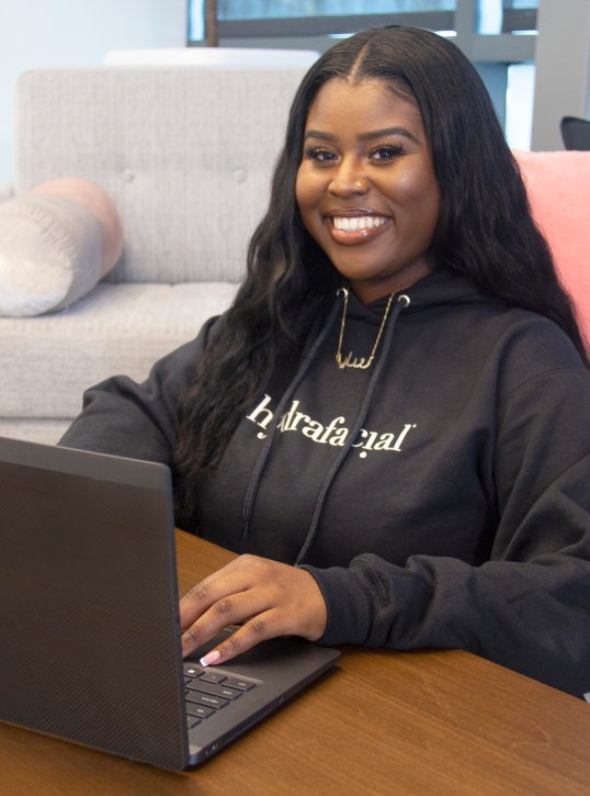 Smiling woman wearing HydraFacial hoodie working on a laptop